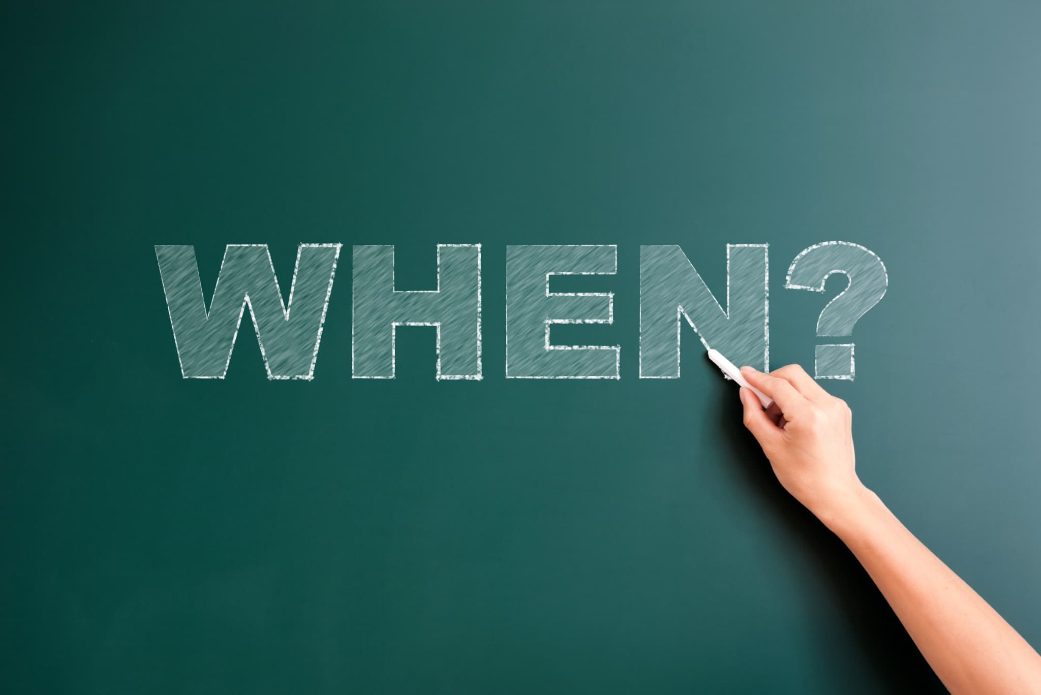歯科医院で検診を受ける頻度はいつかという疑問を示すWHEN？の文字を黒板にチョークで書いている手